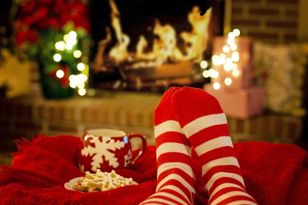 Personne détendue devant un foyer portant des chaussettes rouges et blanches, avec une tasse et des biscuits sur une couverture rouge.