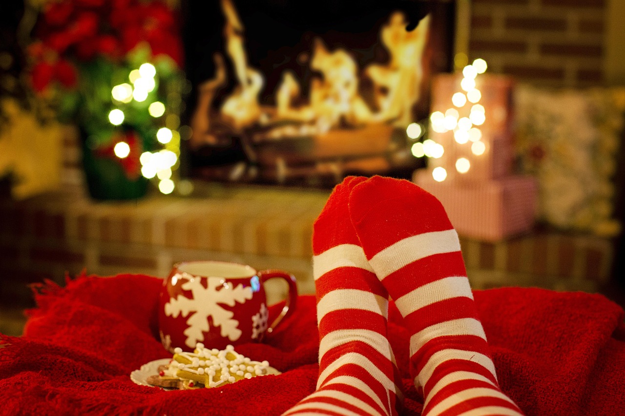 Personne détendue devant un foyer portant des chaussettes rouges et blanches, avec une tasse et des biscuits sur une couverture rouge.
